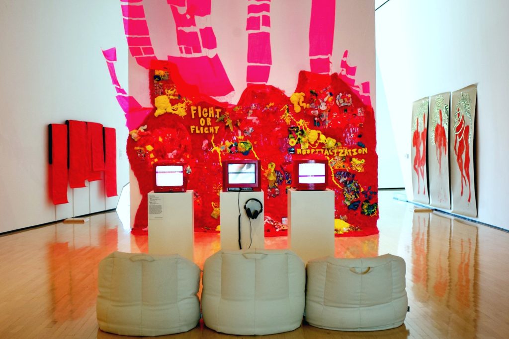 A picture of an art installation. It is a red wall with different toys and bits of yellow material attached together. Three chairs and three TVs sit in front of the red wall. 