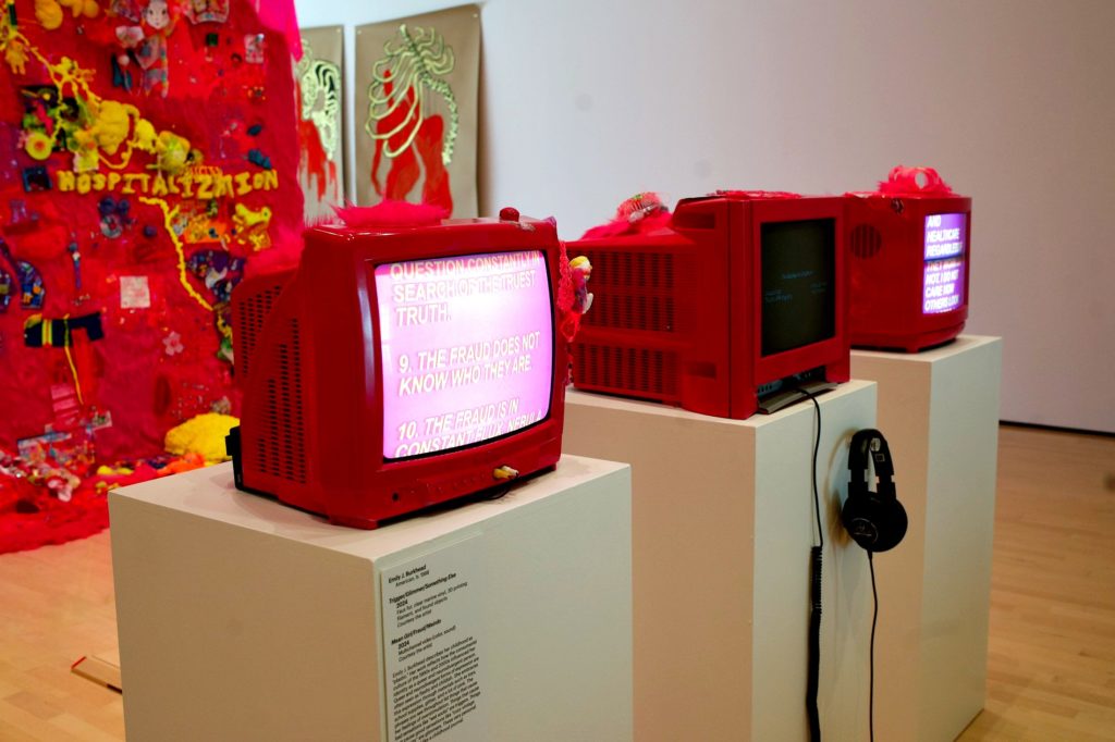 A picture of an art installation. It is a red wall with different toys and bits of yellow material attached together. Three TVs stand in front of it with text scrolling up and down. 