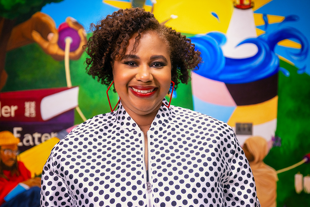 A woman wearing a black and white black polka dot patterned shirt stands in front of a colorful mural.