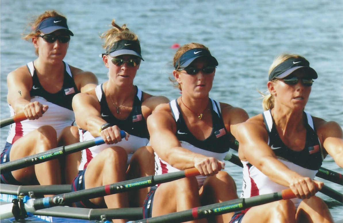 Two-time Olympic rower Kelly Salchow MacArthur (third from the left) is part of a group of four people rowing together on a body of water in Athens, Greece. They wear visor hats and sunglasses and it's a sunny day.