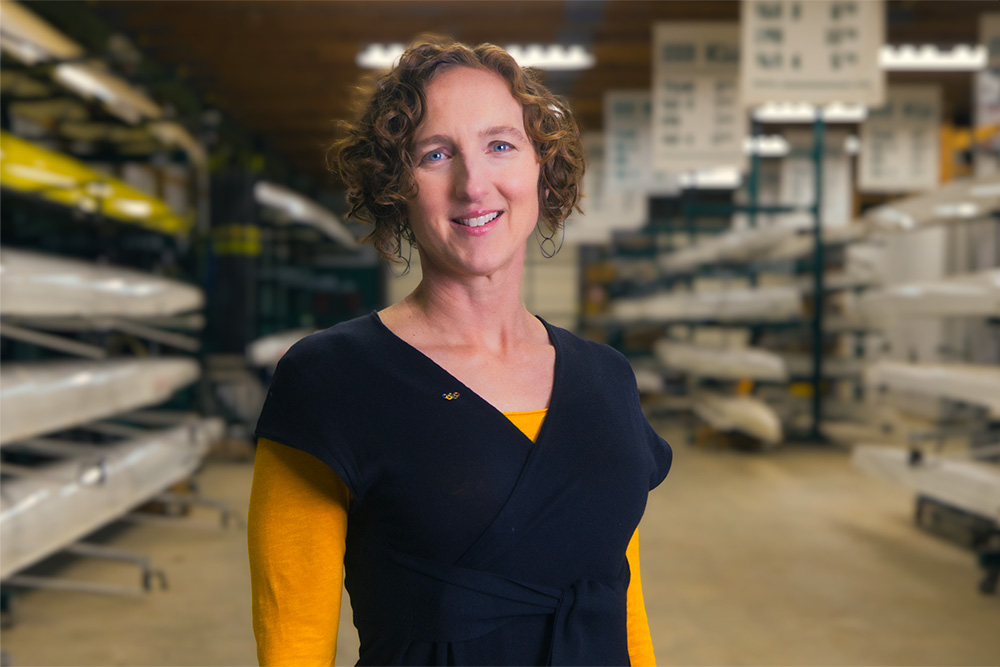 Kelly Salchow MacArthur, a two-time Olympic rower and graphic design professor at Michigan State University, stands in between shelves of rowing boats in the MSU Boathouse. She is wearing a black shirt with yellow sleeves and a pin showing the five Olympic rings.