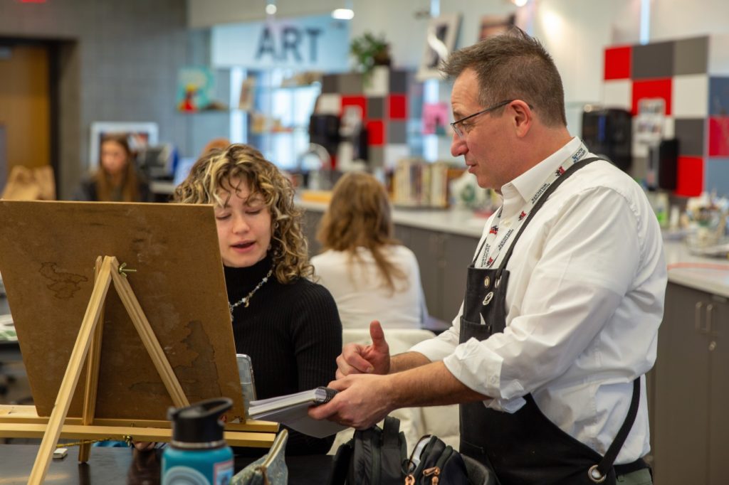 A picture of two people looking at an easel - on the left is a young person with blonde and black hair and a black turtleneck. On the right is a man with short hair, glasses, a white shirt, and an apron. In the background is an art classroom.