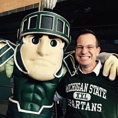Man standing with Sparty, who has his arm around the man, and the man is smiling and wearing glasses and a shirt that says "Michigan State Spartans"