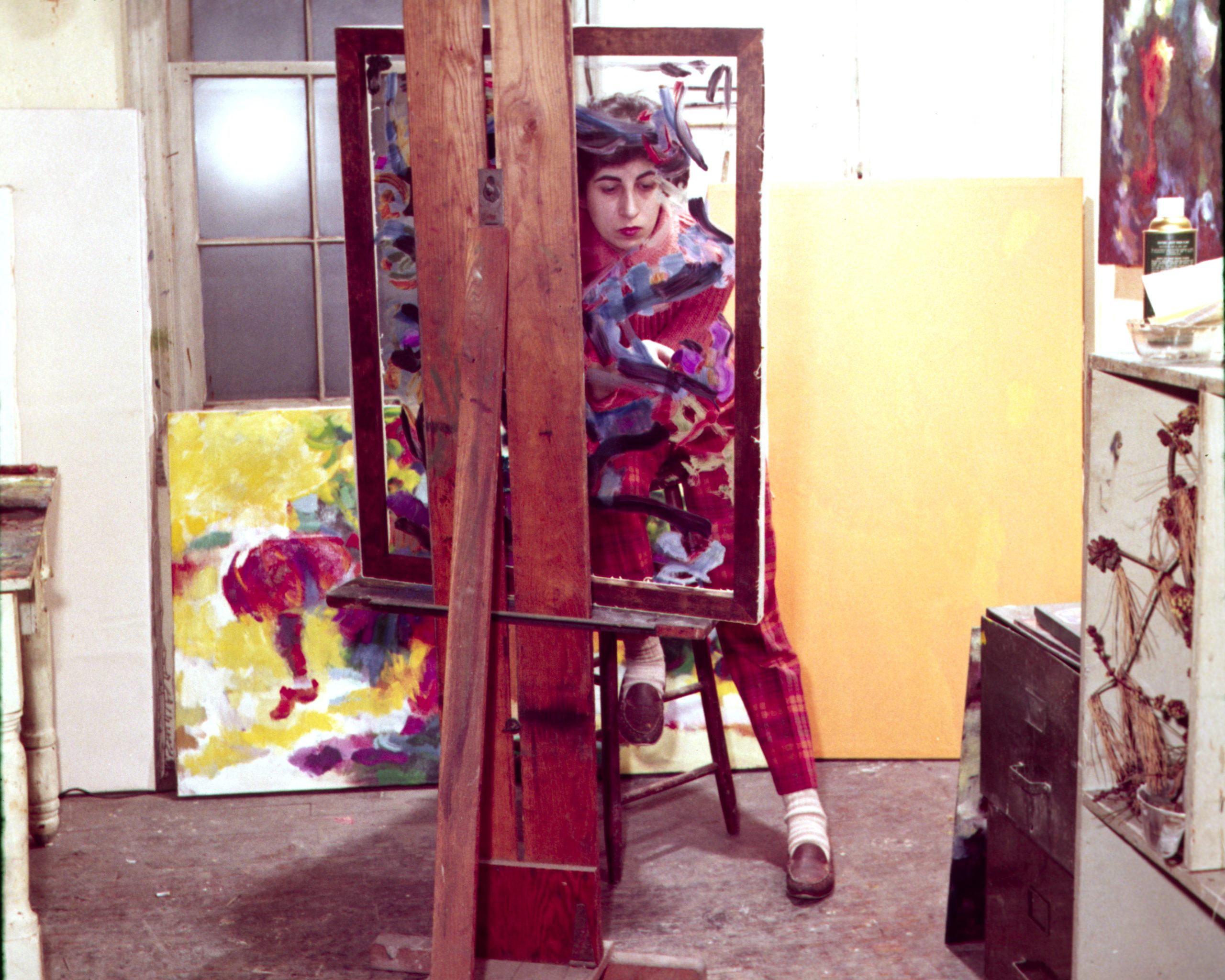 A picture of a woman in an art studio posing in an empty frame, wearing a colorful outfit. 