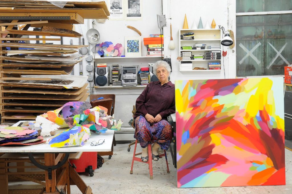 A picture of a woman with short grey hair in an art studio posing next to a large multicolored painting. 