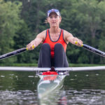 Kelly Salchow MacArthur, a two-time Olympic rower and graphic design professor at Michigan State University, rowing in her boat on a body of water with green trees in the background.