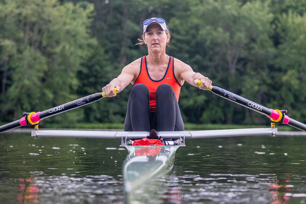 Kelly Salchow MacArthur, a two-time Olympic rower and graphic design professor at Michigan State University, rowing in her boat on a body of water with green trees in the background.