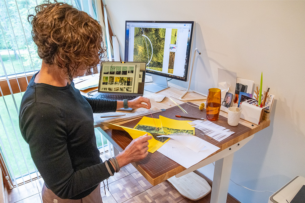 Kelly Salchow MacArthur, a two-time Olympic rower and graphic design professor at Michigan State University, works on graphic design projects at a standing desk with a laptop and larger monitor.