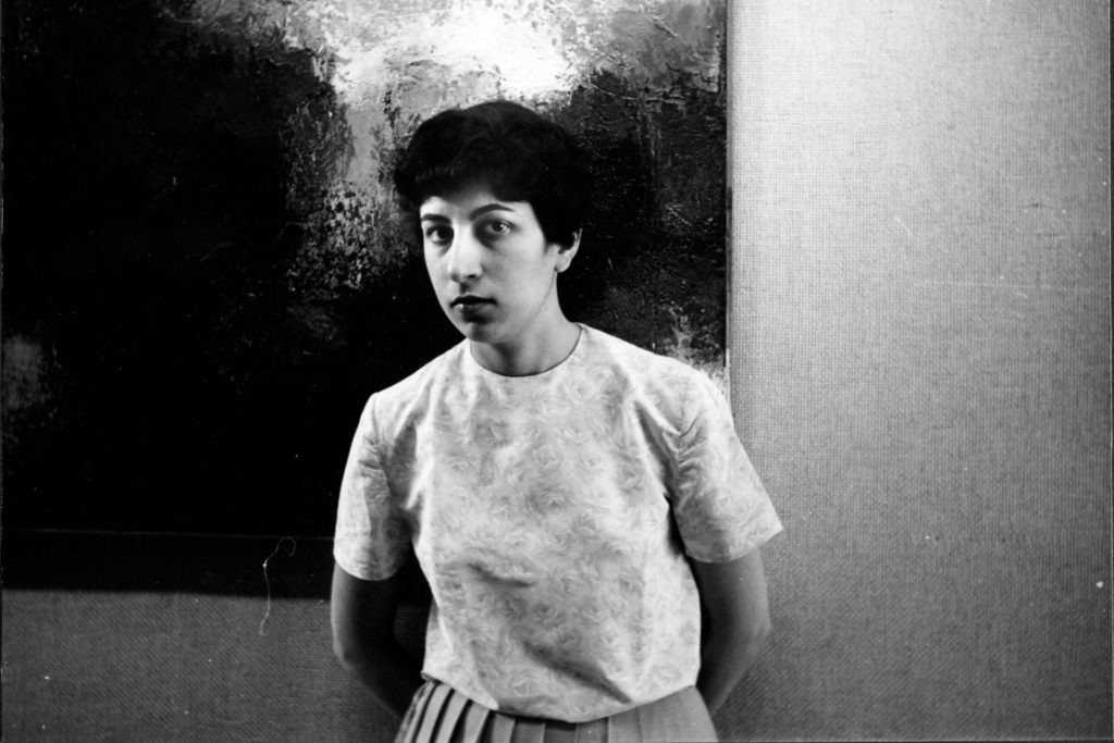 A picture of a woman in black and white. She has short black hair and is posed in front of a painting. 