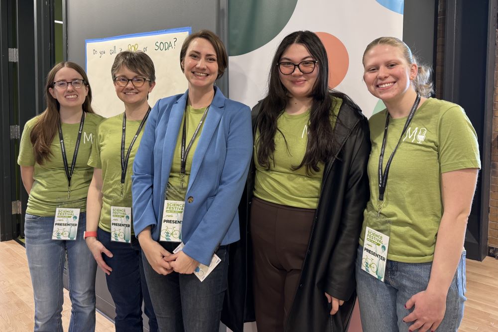 Five people stand in a row in front of a poster on a wall that says: “Do you call it pop or soda?” The people are wearing green shirts and lanyards with nametags that say: “MSU Science Festival Presenter.”