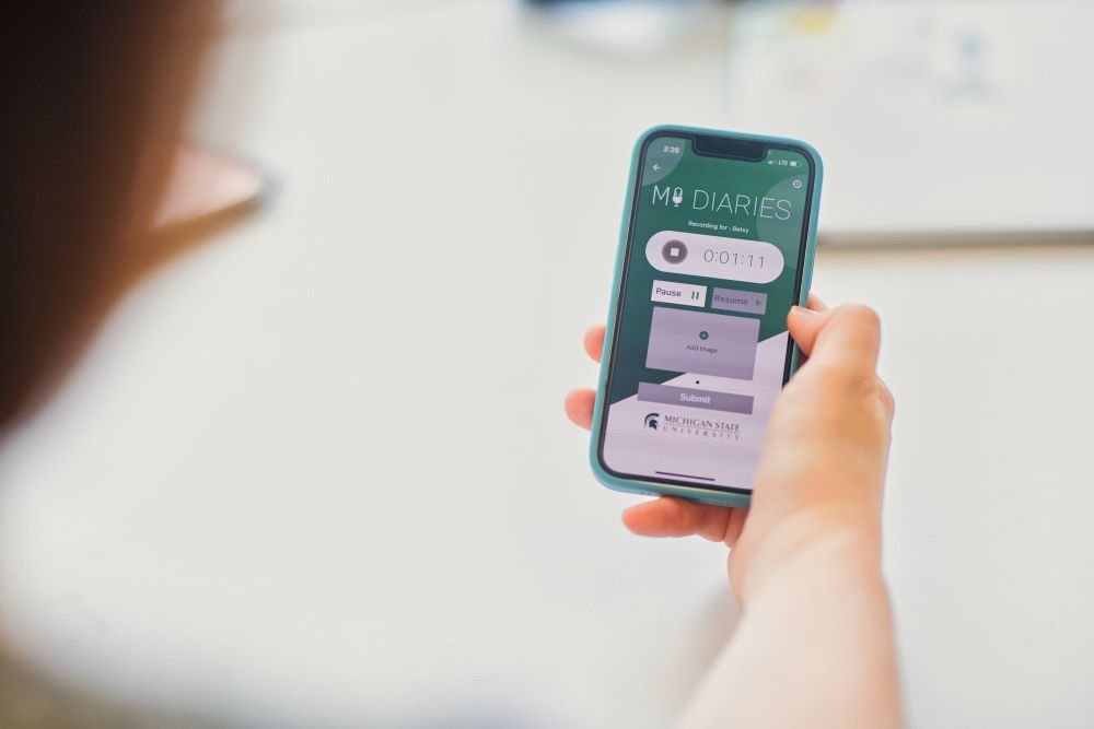 A person holds a mobile photo. On the screen of the phone is a mobile app called MI Diaries that shows a recording bar. At the bottom it says: Michigan State University.
