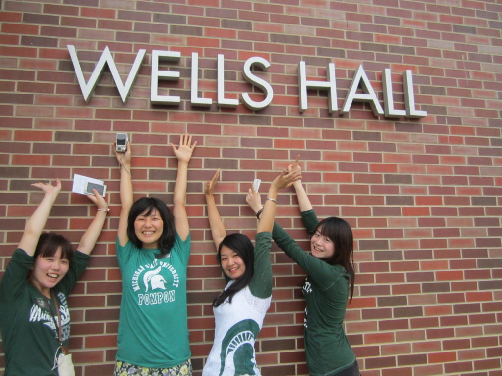 Smiling English Language Center students with arms raised under the Wells Hall building sign