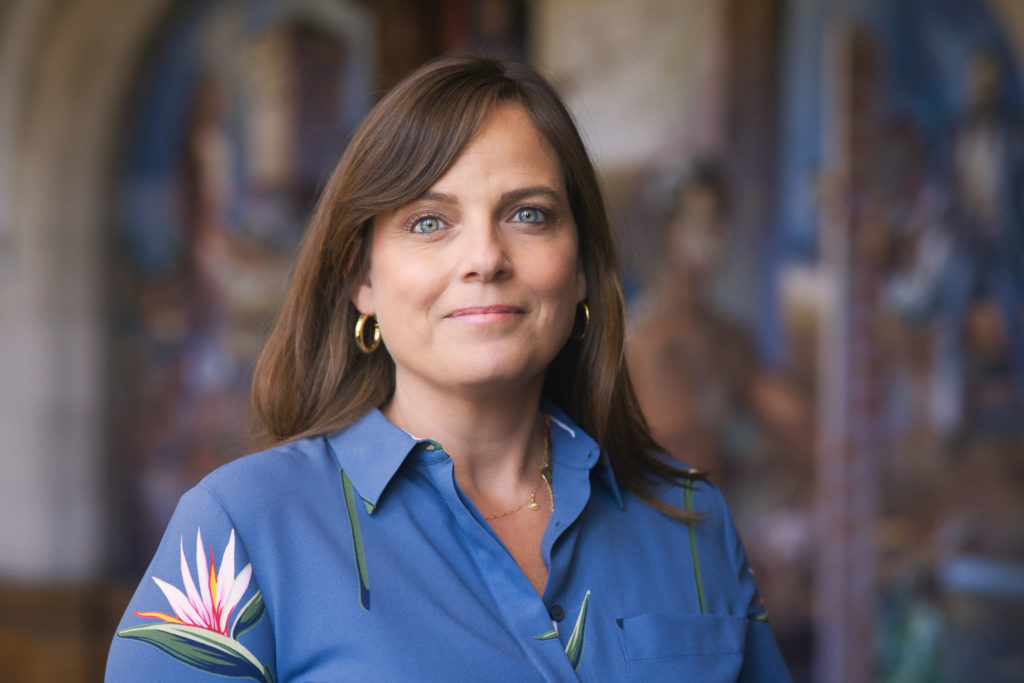 A picture of a woman with shoulder-length brown hair and blue eyes, wearing a blue shirt with a pink flower on one of the arms.