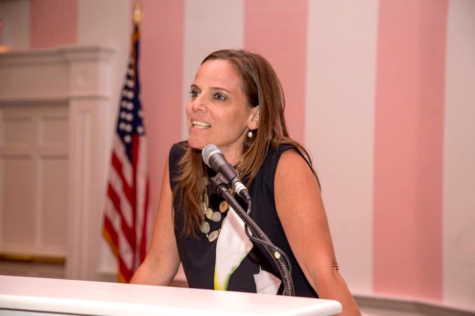 Woman standing at a podium with a microphone and the American flag in the background. 