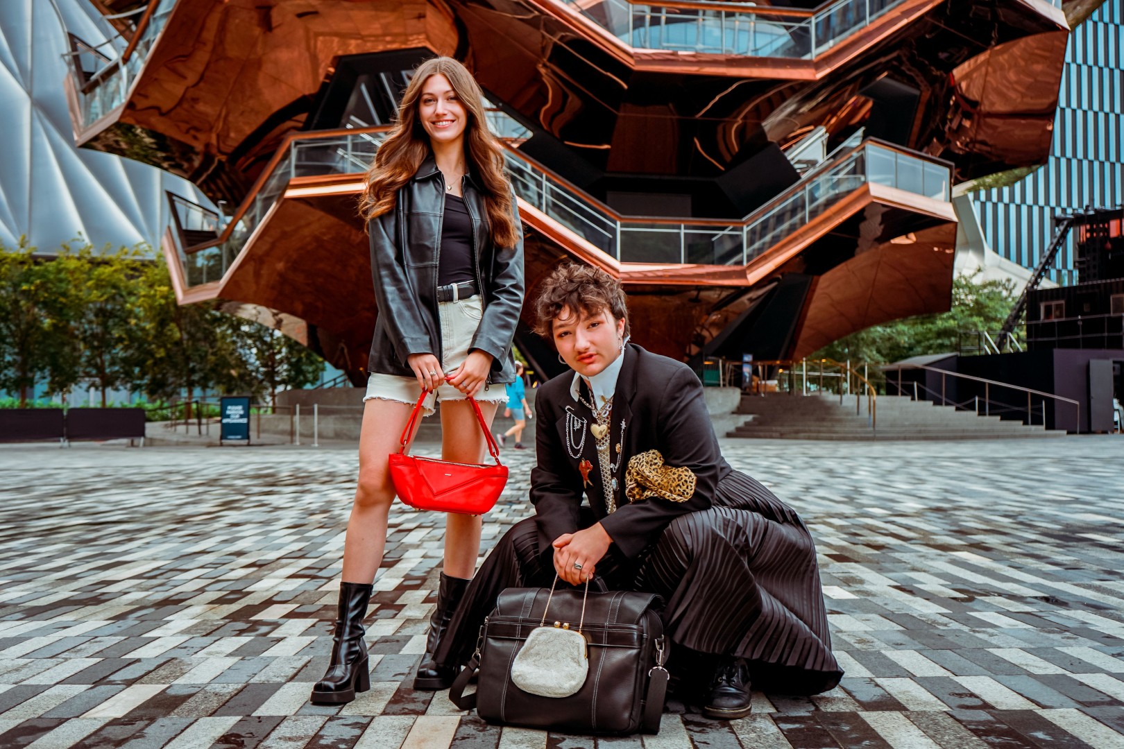 A photo of two people standing in front of an architectural display. The woman on the left has long brown wears a black leather jacket and carries a red purse; the man on the right has short, choppy hair and wears a suit coat covered in pins and a dress.