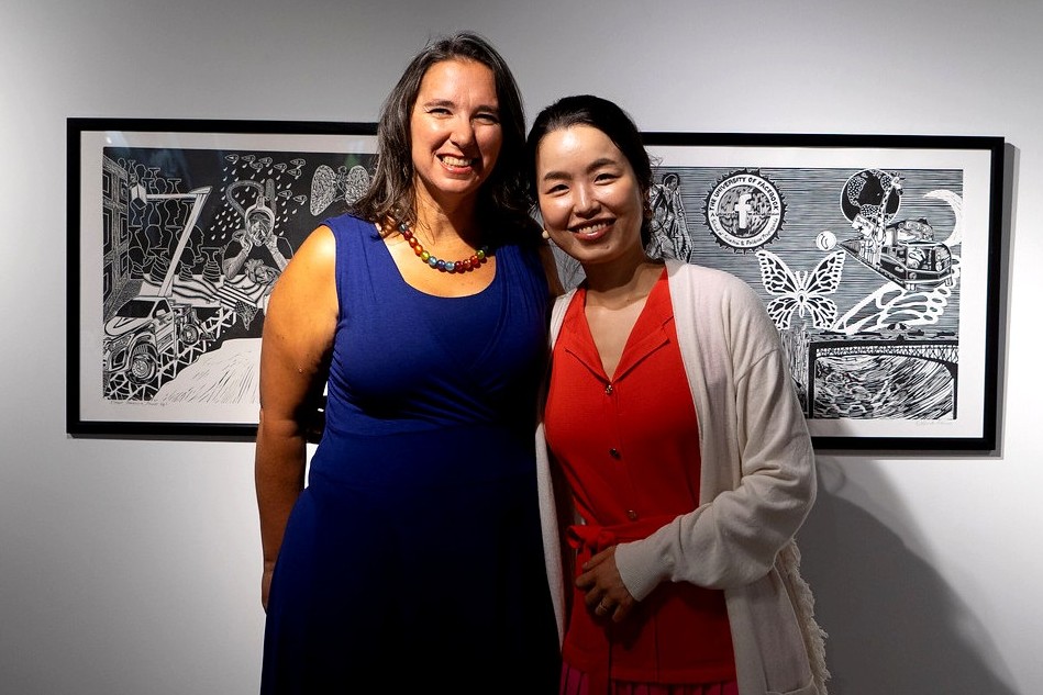 A picture of two people side by side. The person on the left wears a blue dress with a necklace of multi-colored peals; the person on the right wears a red dress with a white cardigan. Behind them is a black and white work of art on a white wall. 