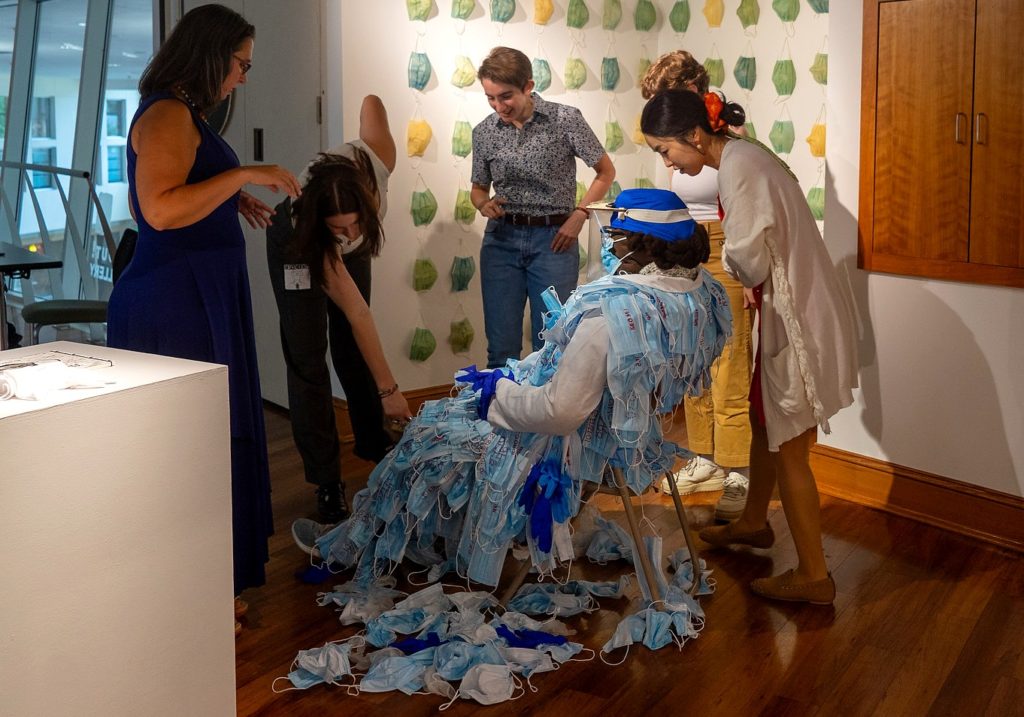 A picture of a group of people standing around someone on a chair, covered in blue cloth masks. On the wall behind them, more masks hang in orderly rows.