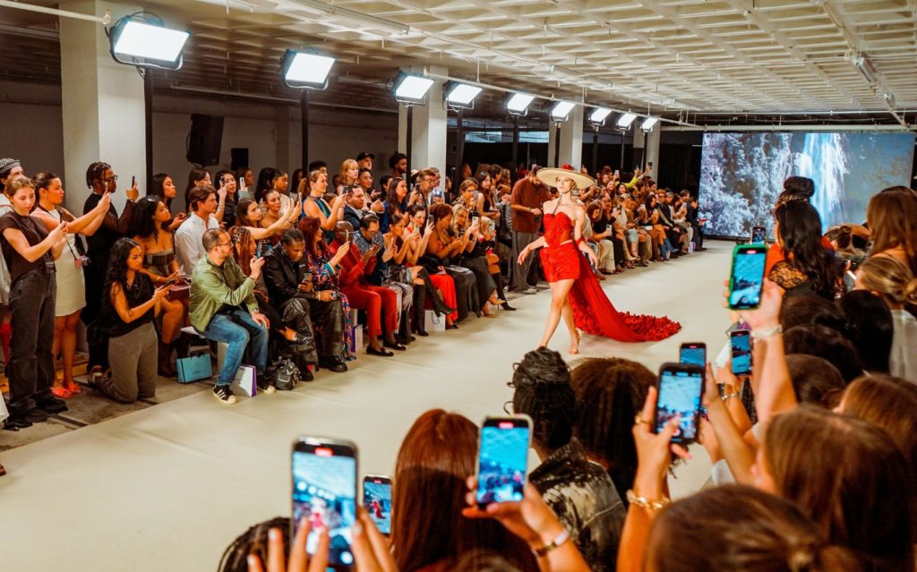 A picture of a fashion show - a model in a red dress and tan hat walks down the runway in the center of the frame, as crowds of people on either side take photos and watch.