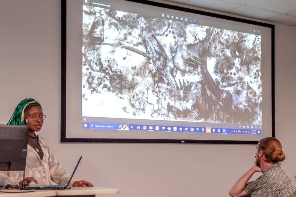 woman standing at a podium with a large screen next to her that is showing a piece of black and white art work.