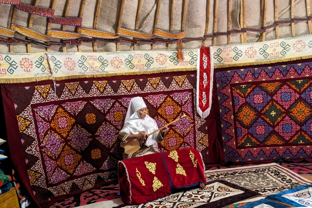 A picture of a person in white and brown ceremonial clothing sitting in front of a wall of multicolored tapestries, featuring patters in red, pink, yellow, orange, blue, and green. 