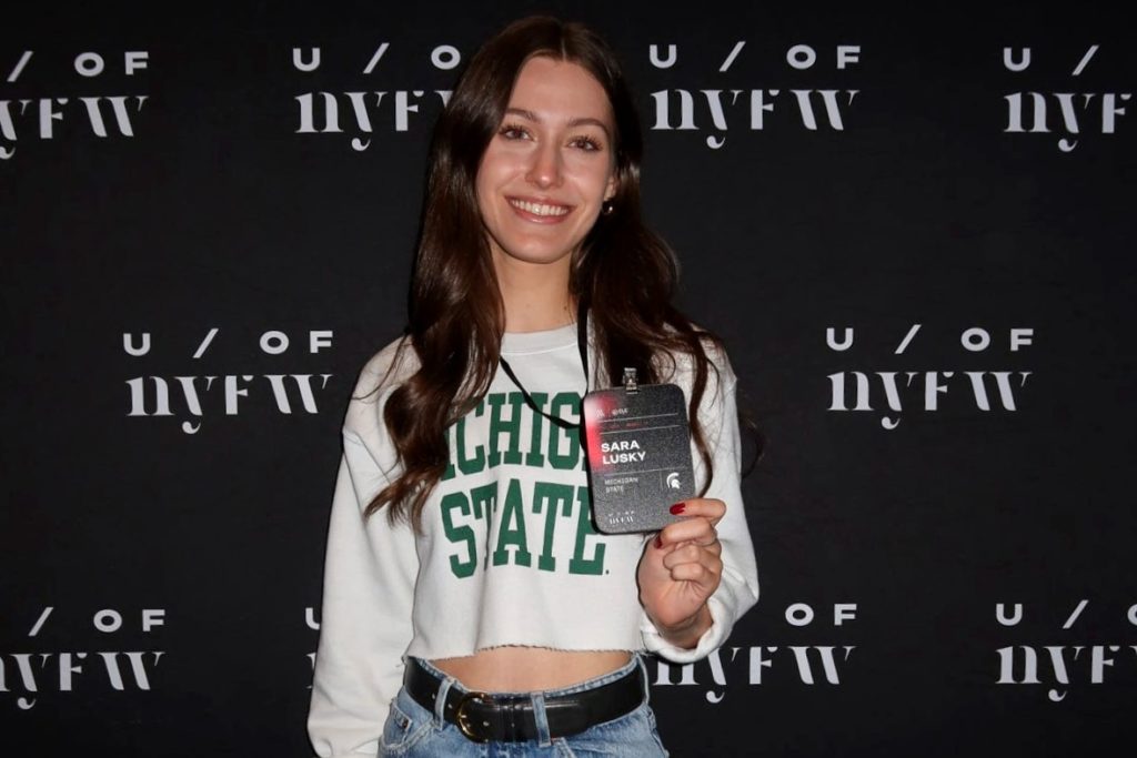 A picture of a woman with long brown hair in a white sweater with Michigan State written on it posing in front of a black background that reads "U / of NYFW"