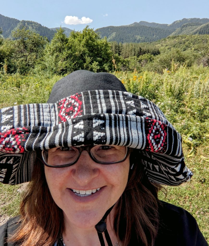 A picture of a woman with a large black hat with red and white embroidery standing in a forested area. She also has glasses and wears a black shirt.