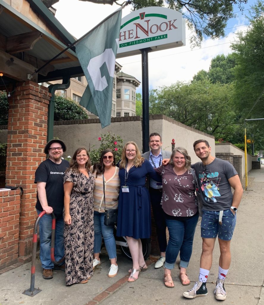 A picture of seven people posing outside a brick building. A green and white flag flies above them, near the top of the frame. In the background are white walls and green trees.