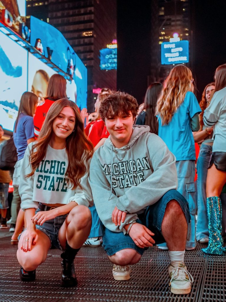 A picture of two people posing in a crowd with neon advertisements in the background. The woman on the left has long brown hair and wears a white sweater with Michigan State written on it. The man on the right has short, messy hair, and wears a grey sweatshirt with Michigan State written on it. 