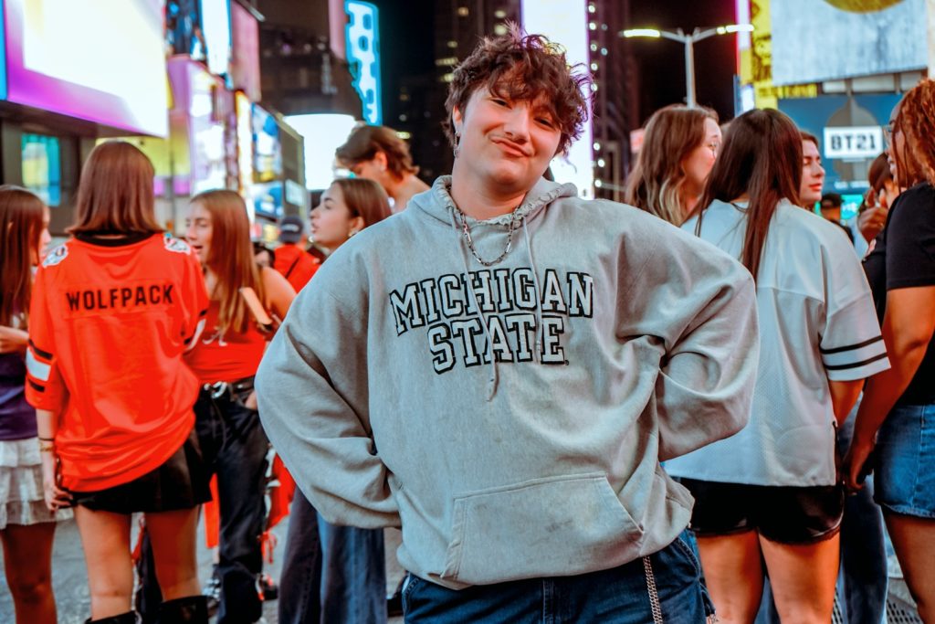 A picture of a man in a crowd, with advertisements and neon lights in the background. He has short, messy hair and wears a grey sweatshirt with Michigan State written on it. 