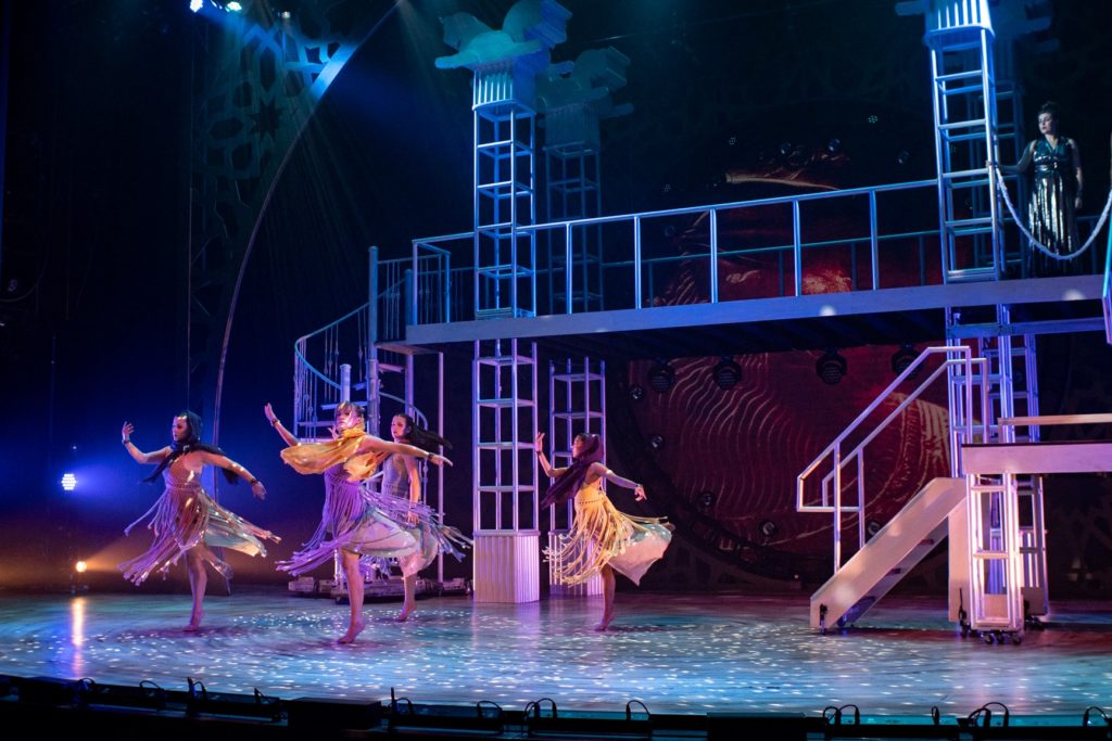 A group of dancers in flowing, fringed costumes performs on a stage illuminated by blue and purple lighting. The set is multi-level with tall columns and spiral staircases.