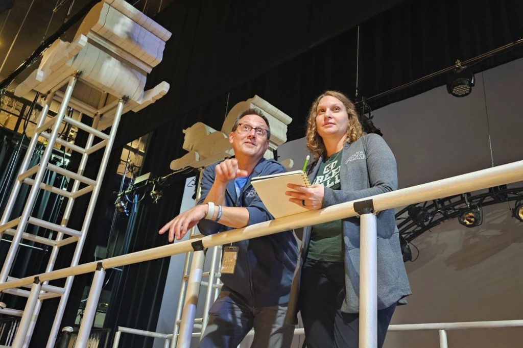 Two people standing on a set platform with stage lighting overhead. The man on the left gestures while the woman on the right holds a notepad. The set features large classical column pieces and ladders in the background.