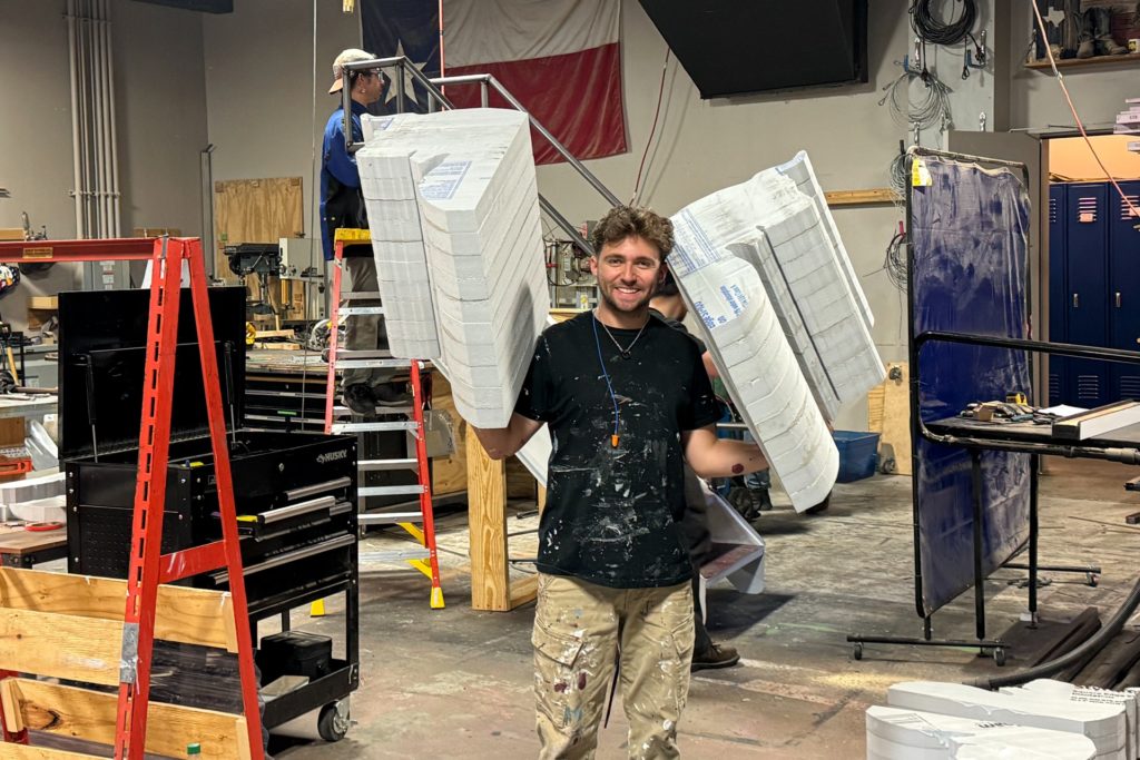 A smiling man in a paint-splattered shirt holds two large, foam architectural pieces resembling classical columns in each arm. The workshop behind him has tools, ladders, and equipment scattered around, with another person working in the background