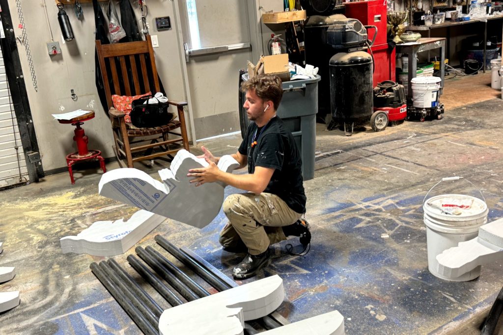 A man kneels on the floor of a workshop, working on a large foam piece shaped like part of a classical column. Surrounding him are additional foam pieces, buckets of paint, and various tools.