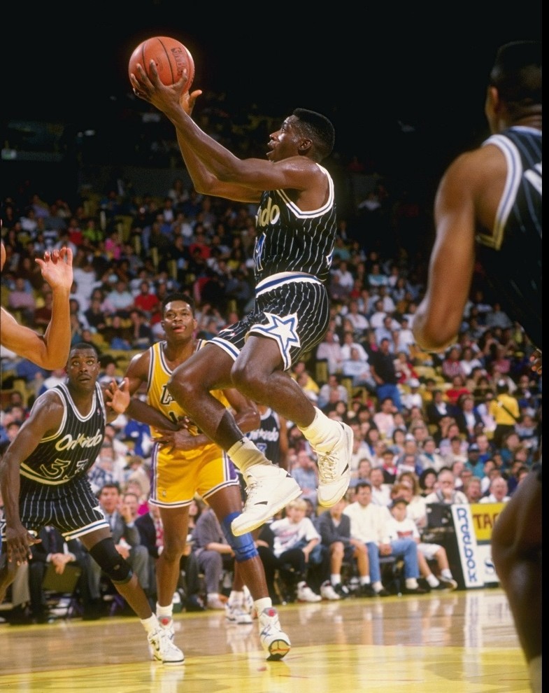 A basketball player from the Orlando Magic attempts a layup during a game against the Los Angeles Lakers, with other players and a packed arena visible in the background.