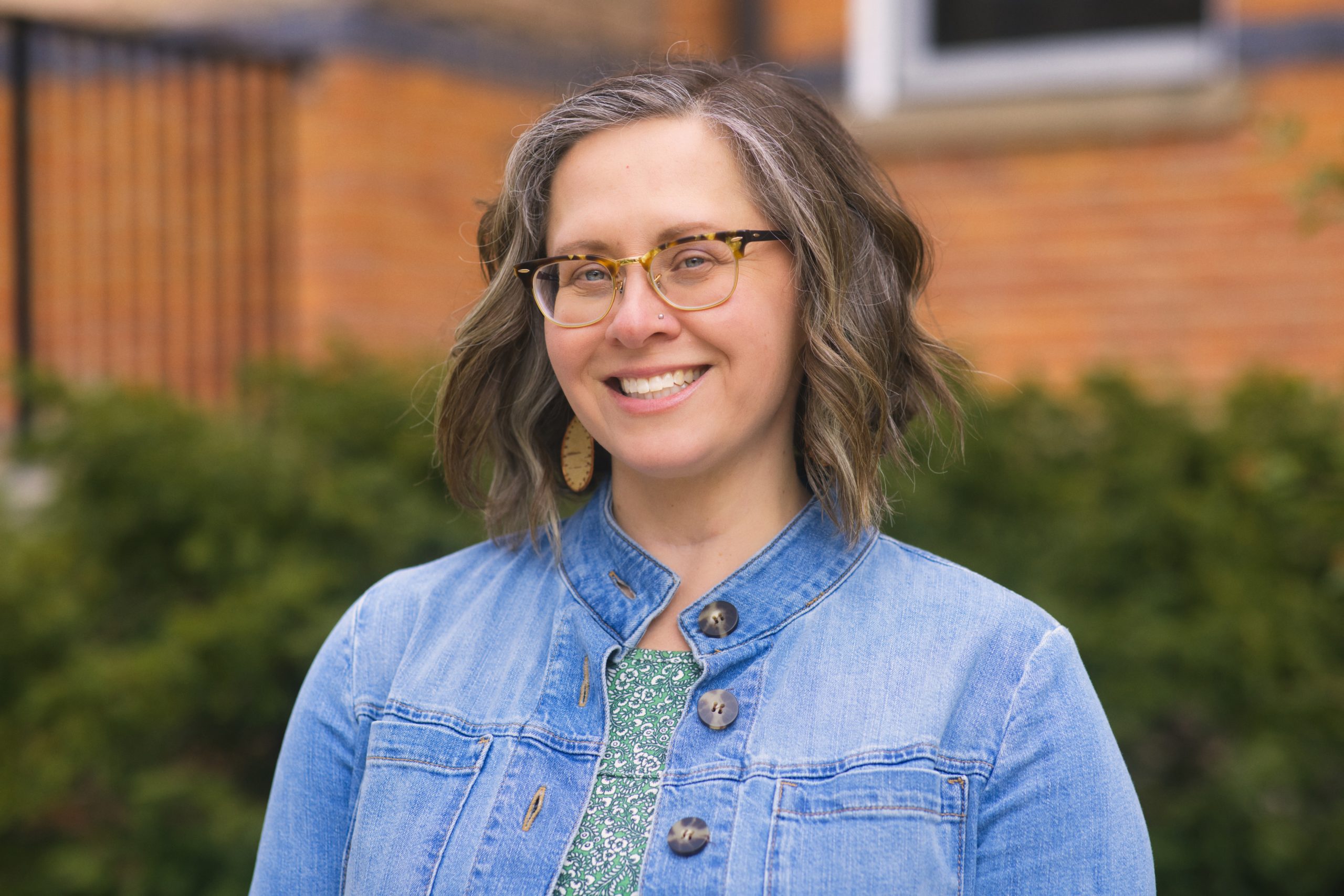 A headshot photo of a woman with short, shoulder-length hair, smiling and wearing glasses and a jean jacket.