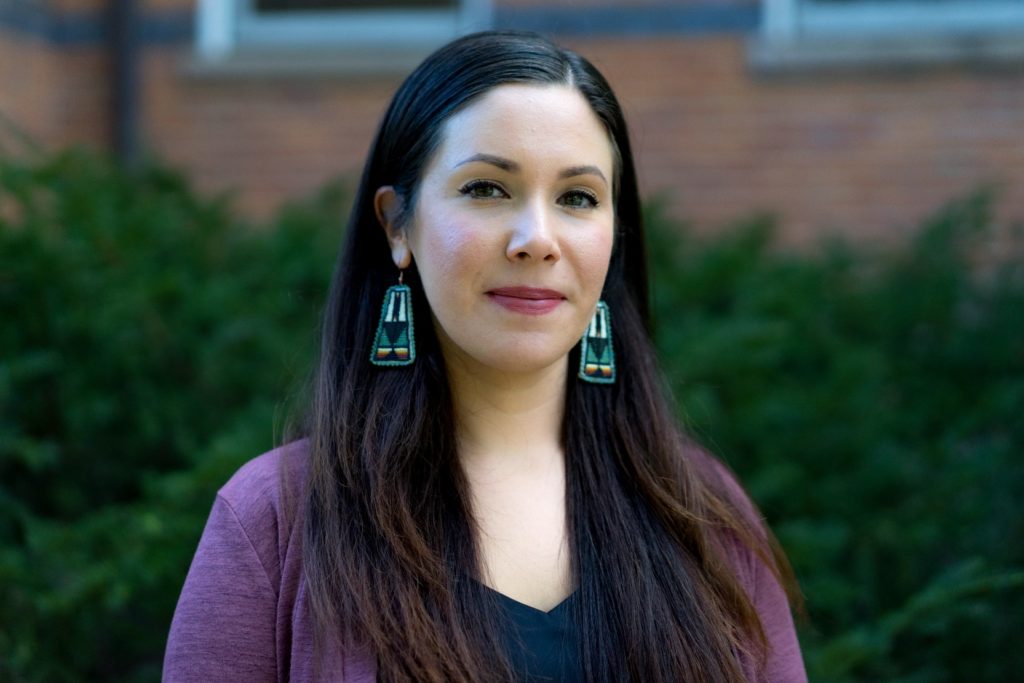 A picture of  woman with long, dark brown hair standing outdoors. She is wearing a dark top and a purple cardigan, with green, orange, and white beaded geometric earrings.