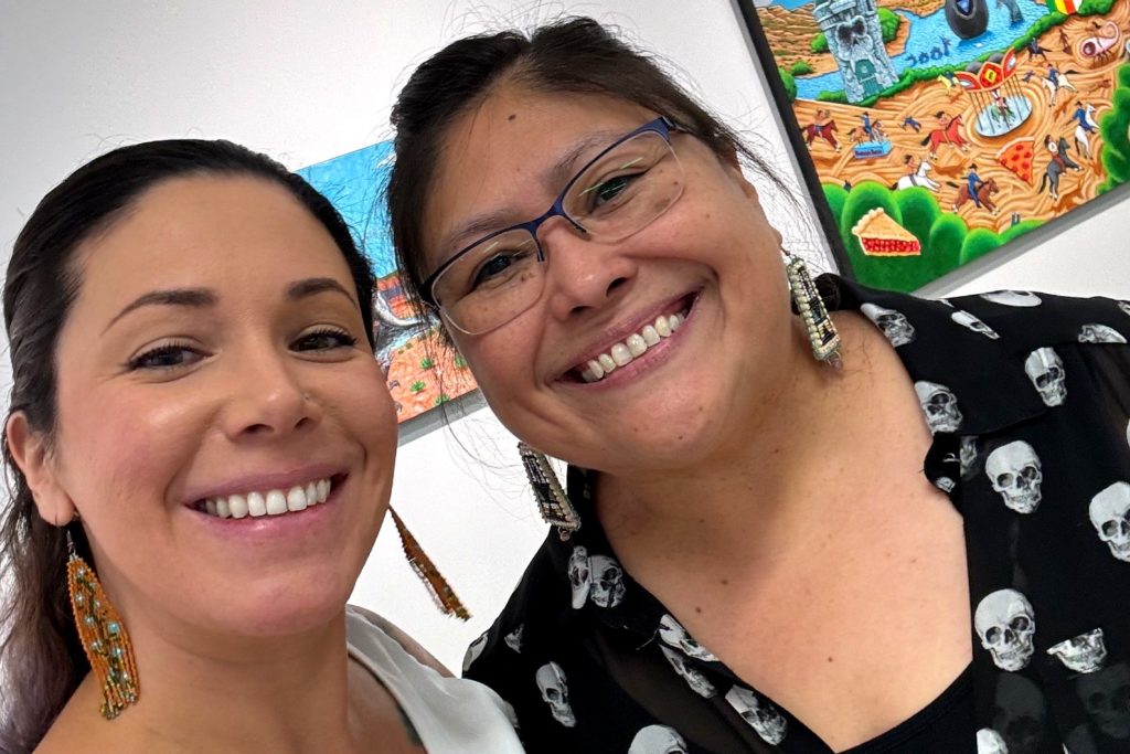 A picture of two women smiling at the camera in an art gallery. The woman on the left is wearing long, beaded earrings and a white top; the woman on the right is wearing glasses, beaded earrings, and a black blouse with a skull pattern. Colorful artwork is visible on the wall in the background.