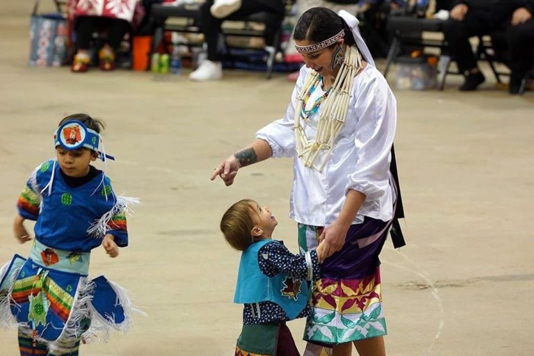 A picture of a woman in a white blouse with beaded accessories smiling and interacting with a young child wearing a blue vest. Another young child, dressed in bright blue regalia and a headband, dances nearby. Chairs and spectators are visible in the background.