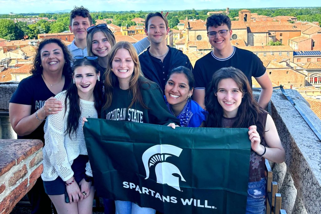 A group of nine individuals posing with a green flag with a Spartan helmet on it, above the text "Spartans Will." The background is a older European city with tan brick houses. 