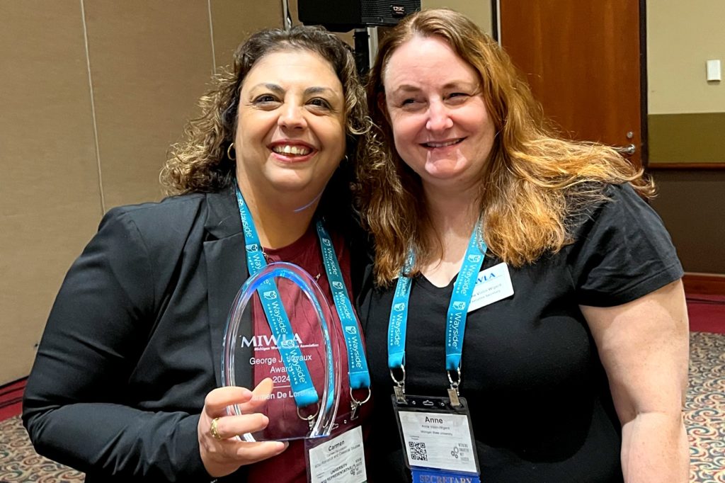 A picture of two women. The woman on the left has long brown hair and wears a black suit jacket over a red top. She holds a glass award in her right hand that reads "MIWLA Georges J. Joyaux Post-Secondary Educator Award." The woman on the right wears a black top and has long blonde hair. Both women wear blue lanyards. 