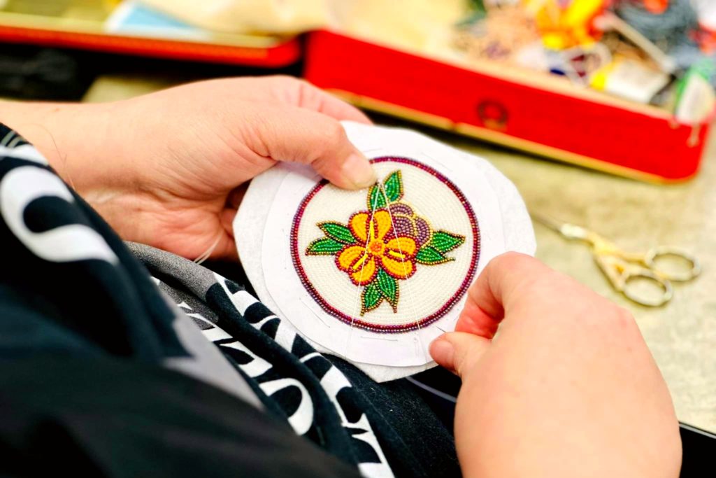 A picture of pair of hands carefully holding a circular piece of fabric featuring an  intricate design of a yellow and purple flower with green leaves, surrounded by a red beaded border.