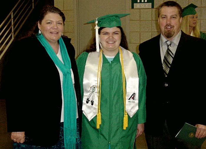 Three people standing next to each other the peron on the left is a woman wearing a black suit. The person in the middle is a woman wearing a green graduation cap and gown, and the person on the right is a man wearing a black suit. 