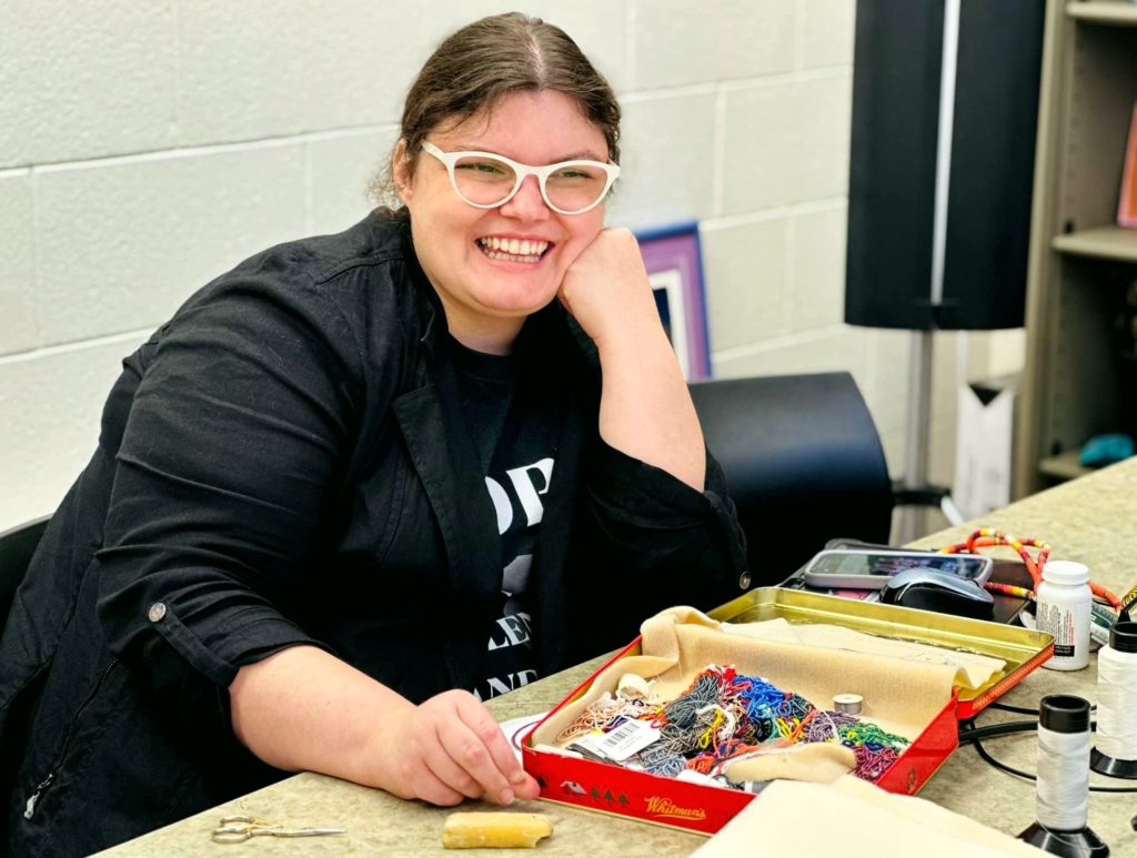 A picture of a woman wearing glasses and a black jacket siting at a table, smiling at the camera. On the table are colorful threads, beads, and sewing tools organized in a red tin box. 