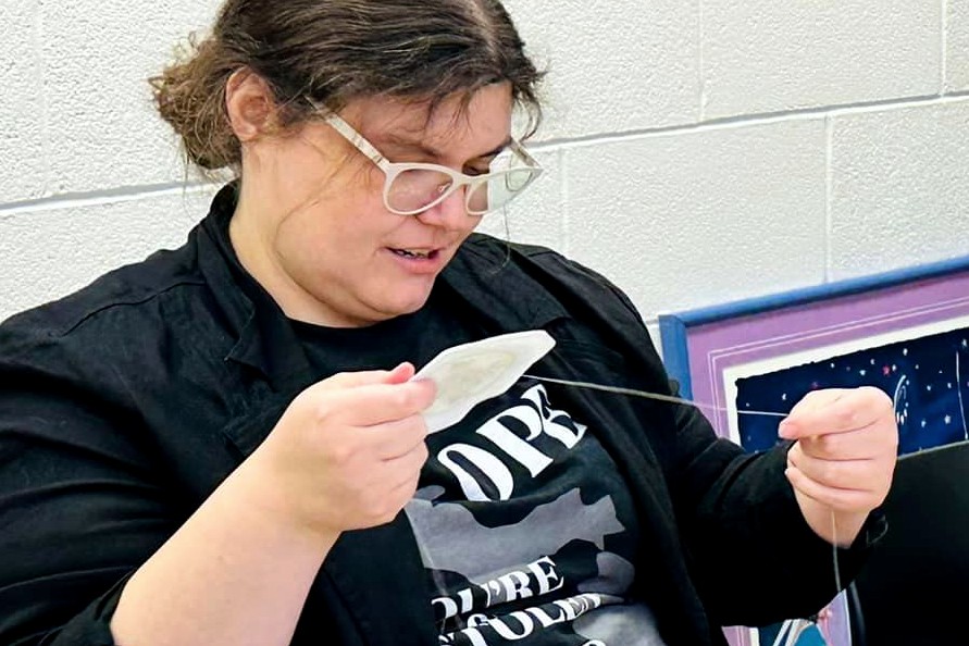 A picture of woman wearing glasses and a black jacket sewing beadwork. She hold a needle and thread in one hand and a circular fabric piece in the other. A white cinderblock wall is visible in the background