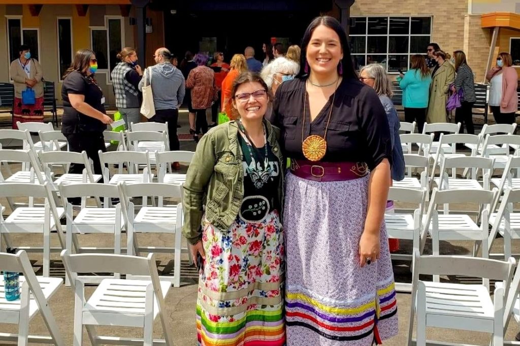 Two women standing next to each other in an area with a lot of white folding chairs and a crowd of people can be seen behind them. The women are smiling and wearing bright-colored skirts and Native American knecklaces. 