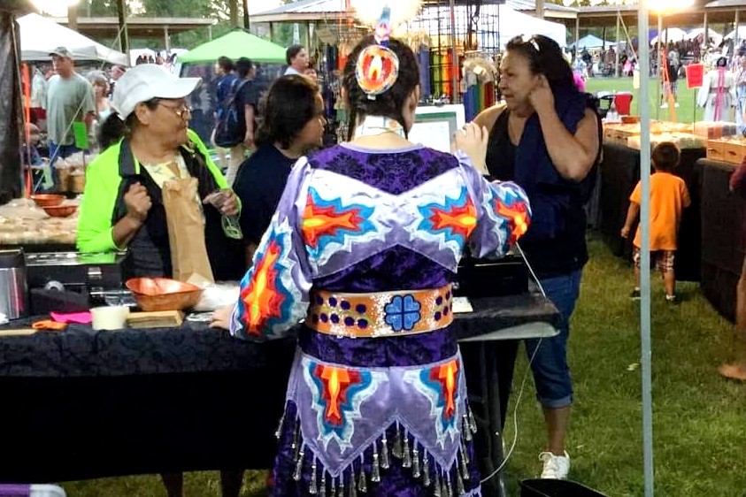  woman wearing a purple Native American dre with her back to the camera. She is looking at three other women who are in plain clothes. They all look to be talking to each other. 