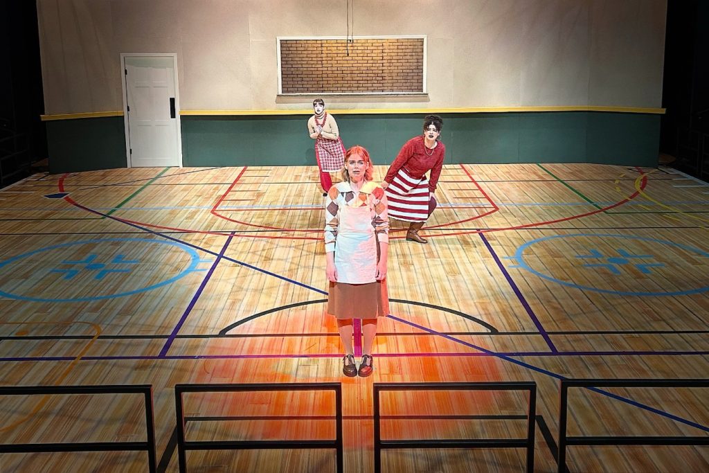 Three female actors on the set of “A Beautiful Day in November on the Banks of the Greatest of the Great Lakes." The three actors are wearing aprons and the set looks like a basketball court. 