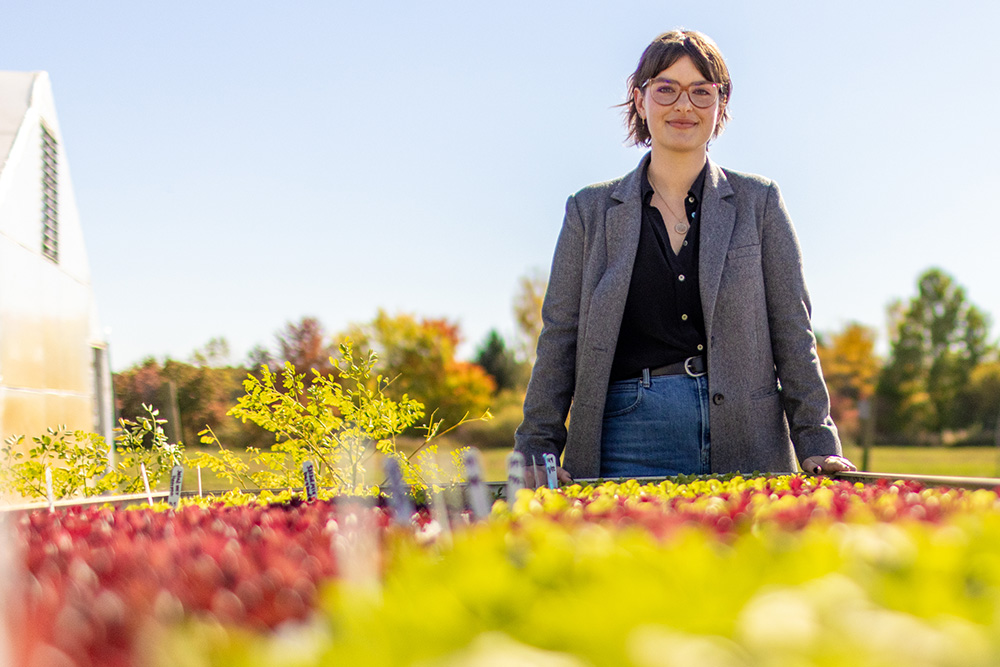 MSU Assistant Professor Megan Dean wearing a blazer and glasses stands next to a raised bed of red and green seedlings. A hoop house is in the background.