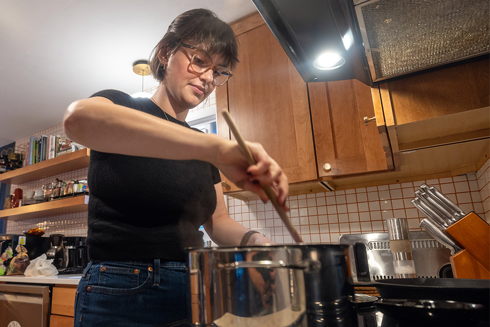 Megan Dean wearing a black shirt and glasses stirs food in a metal pot on a stove in a kitchen setting.