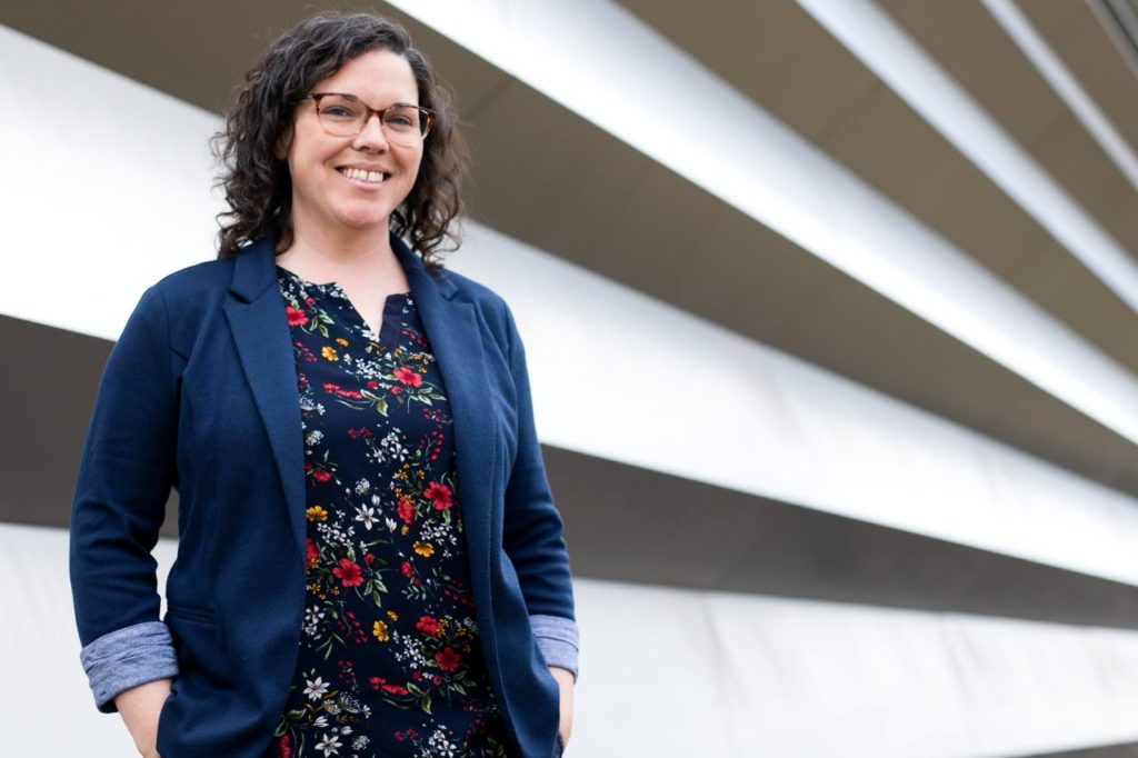 Woman standing with hands in her pockets wearing a blue blazer and a black floral-print top, wearing glasses and brown shoulder-length curly hair.